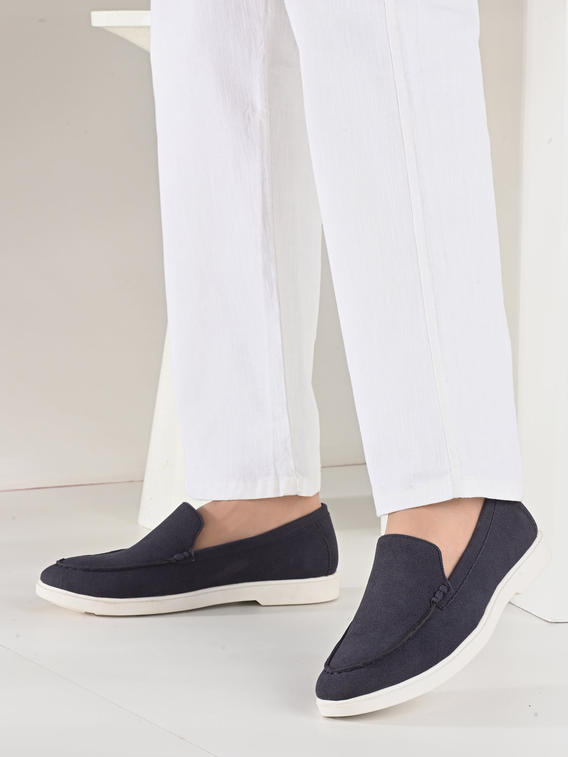 A person wearing white trousers and dark navy slip-on loafers with white soles, shown from the calves down against a clean white studio background.